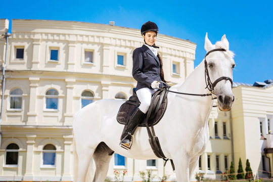 On race track. Elegant gentle woman wearing riding boots riding her white horse on big race track