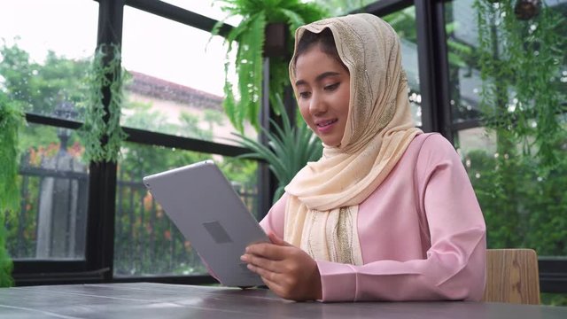 Beautiful young smiling asian muslim woman using tablet sitting in living room at home. Enjoying time at home.