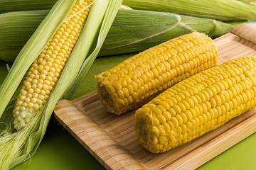 Fresh corn on the cob with leaves and boiled corn in wooden tray on green background