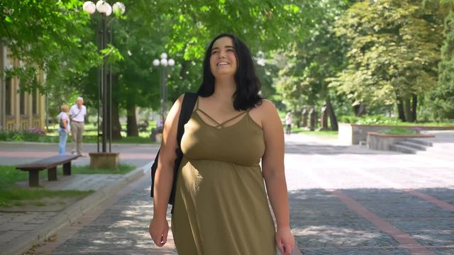Beautiful woman with obesity smiling and looking at camera, standing on street in park, happy and cheerful