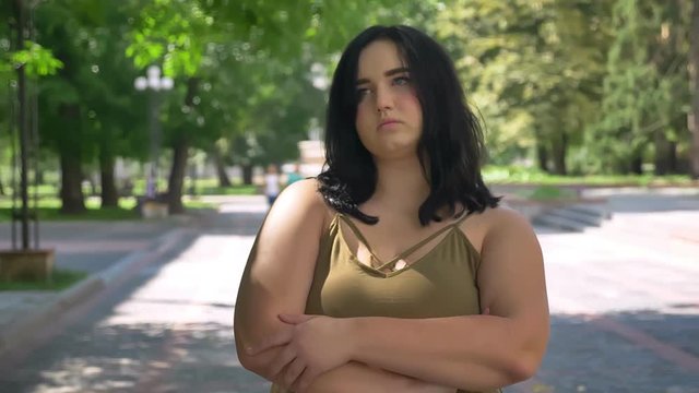 Serious depressed young pretty woman with obesity looking at camera, standing on street in park during sunny day