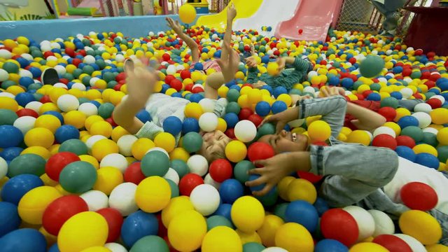 Group of little children of different ethnicities lying in ball pit and smiling when throwing colorful hollow plastic balls