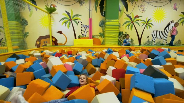 Dolly shot of adorable little girl of elementary school age falling backwards into cube pit and lying in multicolored foam cubes when spending time in indoor playground