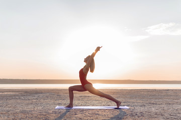Fototapeta premium Girl practicing yoga poses at sunrise. Virabhadrasana pose.