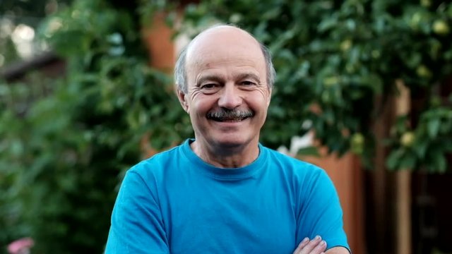 Charming handsome senior man in casual blue shirt keeping arms crossed and smiling while standing on formal garden. At first he is sad, but after hearing a joke begin to smile