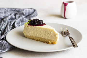 Piece of New York style cheesecake with black currant sauce and rosemary on grey plate on light background.