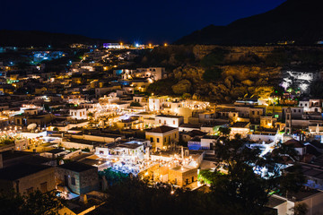 Obraz premium Scenic landscape photo of night Lindos town and castle on Rhodes island, Dodecanese, Greece. Panorama with bright lights, mountains and sea. Famous tourist destination in South Europe