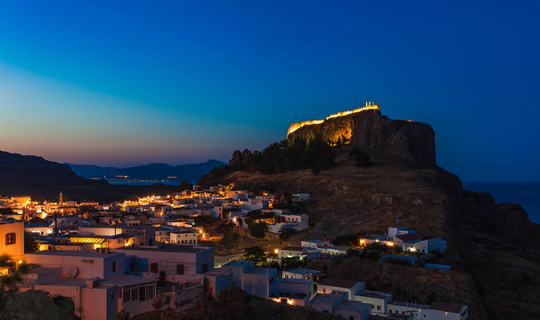 Scenic landscape photo of night Lindos town and castle on Rhodes island, Dodecanese, Greece. Panorama with bright lights, mountains and sea. Famous tourist destination in South Europe
