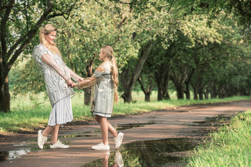 Fototapeta premium beautiful mother and adorable daughter in transparent raincoats holding hands on wet road in green park