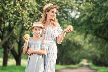 smiling mother and daughter in straw hats looking at appples in hands