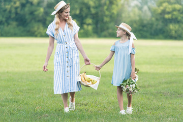 Fototapeta premium beautiful mother and daughter holding wicker basket with fruits and walking on green lawn