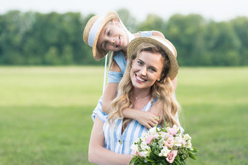 Obraz premium smiling mother and daughter posing in straw hats with bouquet of flowers