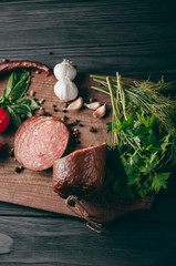 Smoked salami on a black table and a brown board. Sausages with rosemary, garlic and pepper and tomato cherry, dill, parsley.