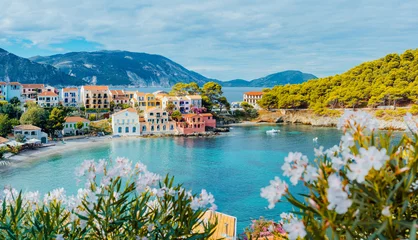 Fotobehang Panoramic view to Assos village in Kefalonia, Greece. Bright white blossom flower in foreground of turquoise colored calm bay of Mediterranean sea and beautiful colorful houses in background © Igor Tichonow