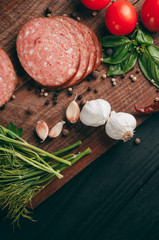 Smoked salami on a black table and a brown board. Sausages with rosemary, garlic and pepper and tomato cherry, dill, parsley.