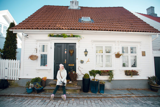 Beautiful young blonde woman in white winter coat sitting on the house porch on sunny day. Cozy house. Architecture. Norway. Stavanger