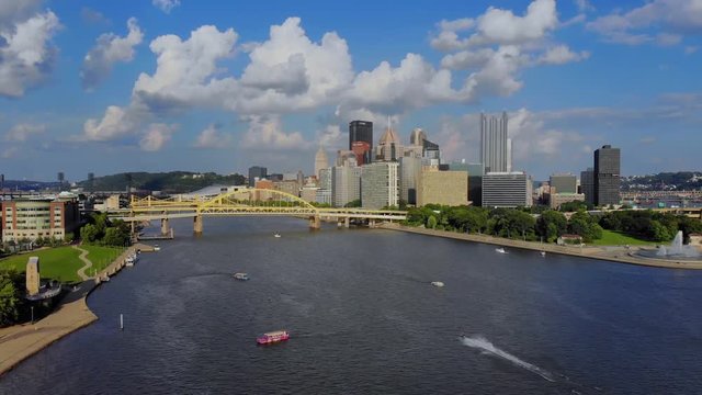 A slow reverse aerial establishing shot of the skyline of Pittsburgh, Pennsylvania on an early summer evening. Ducky tour boats and jet skis in the river below.  	