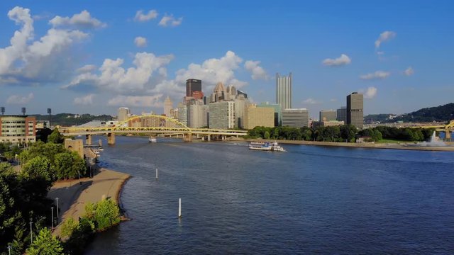 A slow forward aerial establishing wide shot of the skyline of Pittsburgh, Pennsylvania on an early summer evening. Gateway Clipper riverboat carries tourists in the distance.  	