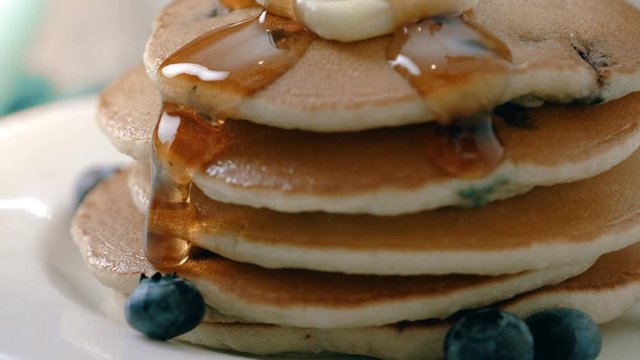 Blueberry pancakes with melting butter and maple syrup being poured and dripping on breakfast plate. 