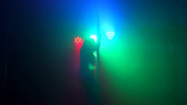 A flexible dancer performs a complex exercise on a pylon in the dark with the light of green, red and blue projectors. Pole dance. Slow motion. Silhouette.