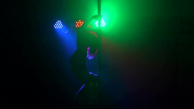 A flexible dancer posing on a pylon in the dark with the light of green, red and blue projectors. Pole dance. Slow motion. Silhouette.