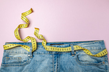 Jeans and yellow measuring tape instead of belt on pink background. Concept of weight loss, diet,...