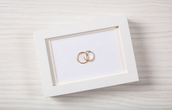 Golden wedding rings on a blank white photo frame, top view, isolated on a white wooden background.
