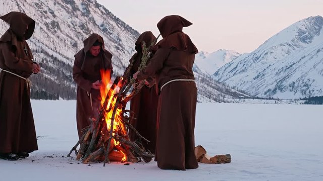 Group of monks in brown cloaks of the order of St. Francis are warming themselves by the fire
