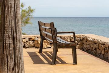Fototapeta na wymiar A bench and a single tree on the promenade in the holiday resort Cala Millor with a beautiful view of Mittelmerr on the Spanish Balearic island Mallorca in front of a radiant blue sky