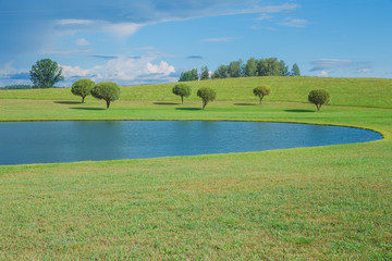 Sunny day, lake and meadow with blue sky. Treeas and clouds.
