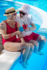 Summer drinks. Beaming mature man and woman having summer drinks while sitting near big outside pool