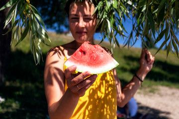 Young beautiful woman laughing happily, smiling and eating a watermelon under tropical leaves of a green tree, in the background a green grass and beach