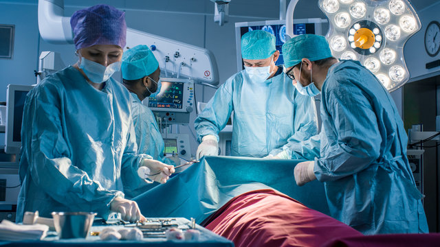 Diverse Team of Professional Surgeons Performing Invasive Surgery on a Patient in the Hospital Operating Room. Nurse Picks Up Instruments, Anesthesiologist Monitors Vitals. Real Modern Hospital.