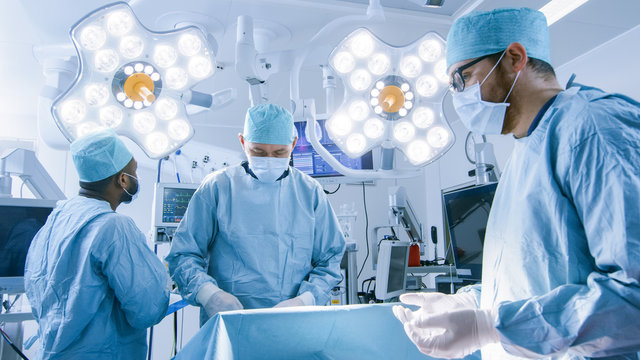 Shot of Diverse Team of Professional Surgeons Performing Invasive Surgery on a Patient in the Hospital Operating Room. Anesthesiologist Monitors Vitals. Modern Hospital.