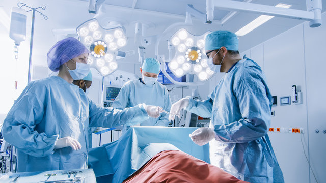 Shot of Diverse Team of Professional Surgeons Performing Invasive Surgery on a Patient in the Hospital Operating Room. Nurse Hands Out Instruments to surgeon,  Anesthesiologist Monitors Vitals.