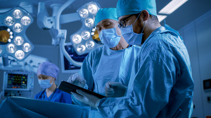 Professional Surgeons and Assistants Talk and Use Digital Tablet Computer while Standing in the Modern Hospital Operating Room. - obrazy, fototapety, plakaty