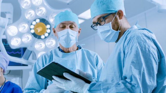Two Professional Surgeons Use Digital Tablet Computer while Standing in the Modern Hospital Operating Room.