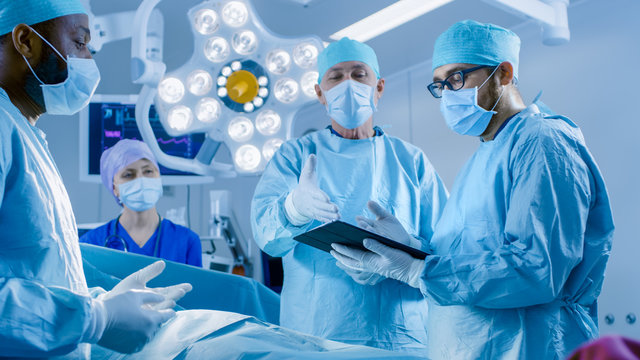 Professional Surgeons and Assistants Talk and Use Digital Tablet Computer During Surgery. They Work in the Modern Hospital Operating Room.