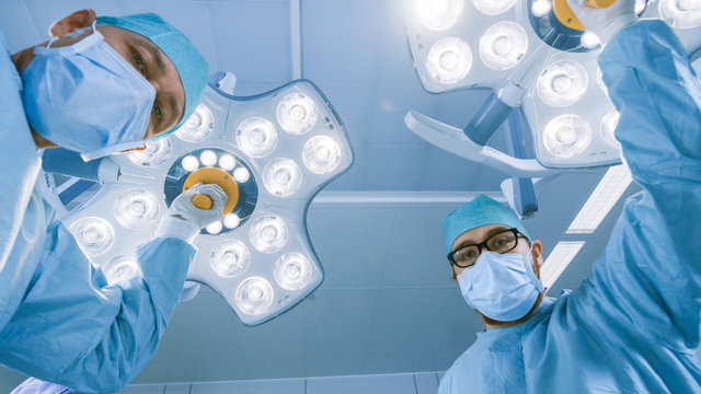 Low Angle Shot POV Patient View: Two Professional Surgeons Turning on Surgery Lights while Bending over Patient.