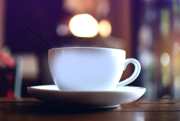 A white cup of hot coffee with a spoon on a dark wooden table in a coffee shop and bokeh green garden background, low key