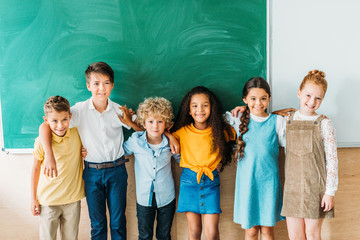 group of multiethnic schoolchildren embracing in front of blank chalkboard