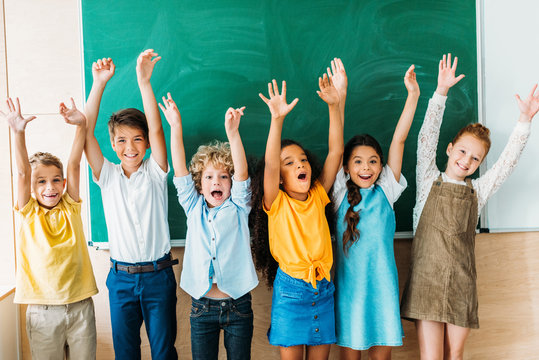 adorable happy schoolchildren with raised hands standing in front of blank chalkboard