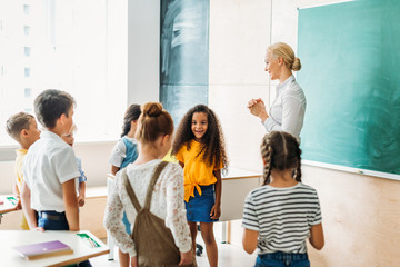 happy multiethnic classmates standing around teacher at classroom