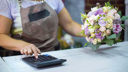 Female florist counting cost of bouquet with calculator in a flower shop. Ribbons, flowers, calculator on working table. shopping, sale, floristry and consumerism concept
