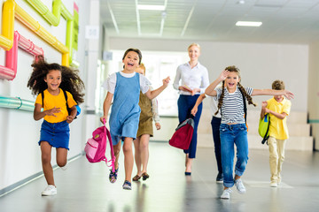 adorable happy schoolchildren running by school corridor together with teacher walking behind