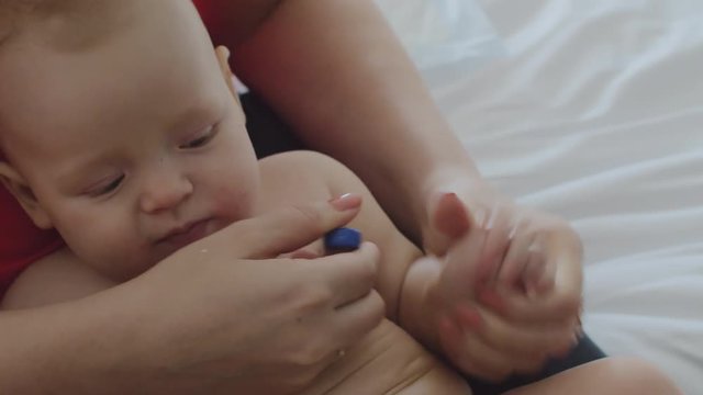 A six months old baby girl with bright grey eyes is lying on a female lap. Her mother is raising her arm and putting a thermometer under it. Then woman is taking the thermometer away, checking it and