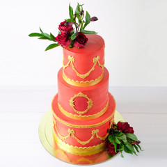 Three-tiered red wedding cream cake decorated with flowers and gold elements on a white wooden table. Picture for a menu or a confectionery catalog.