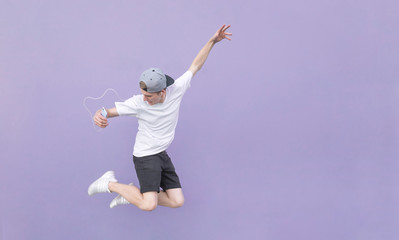 Young man in a white T-shirt and summer clothes listens to music in headphones and jumps on a...