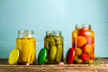 Jars with pickles of courgettes, cucumbers, tomatoes on a blue, wooden background. Stock of food