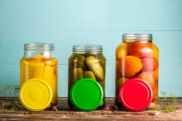 Jars with pickles of courgettes, cucumbers, tomatoes on a blue, wooden background. Copy space